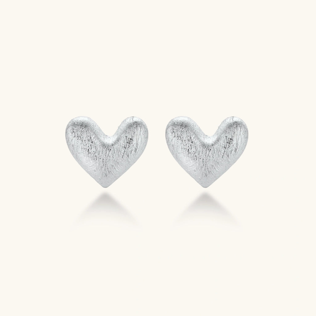 Pair of small heart-shaped stud earrings with brushed metallic finish, showing both silver and gold variants on neutral background.
