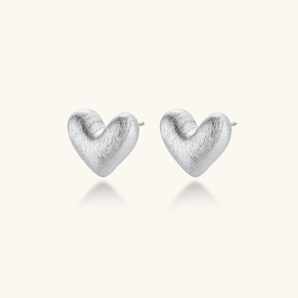 Pair of small heart-shaped stud earrings with brushed metallic finish, showing both silver and gold variants on neutral background.