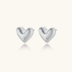Pair of small heart-shaped stud earrings with brushed metallic finish, showing both silver and gold variants on neutral background.
